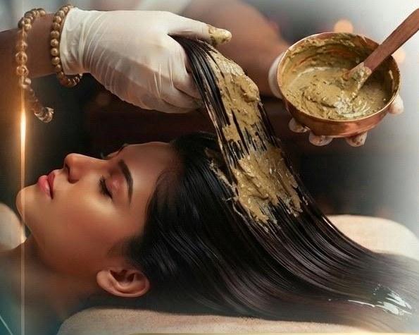 A gloved practitioner working herbal paste through the length of a reclining patient's hair from a copper bowl