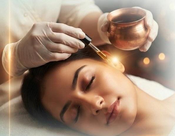 A gloved practitioner holding a copper bowl and dropper above the ear of a reclining patient
